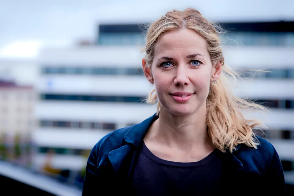 En kvinna med blont hår som står framför en byggnad. Hon kollar in i kameran.