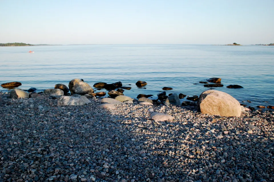 En stor sten som ligger på en strand intill ett vattenområde.